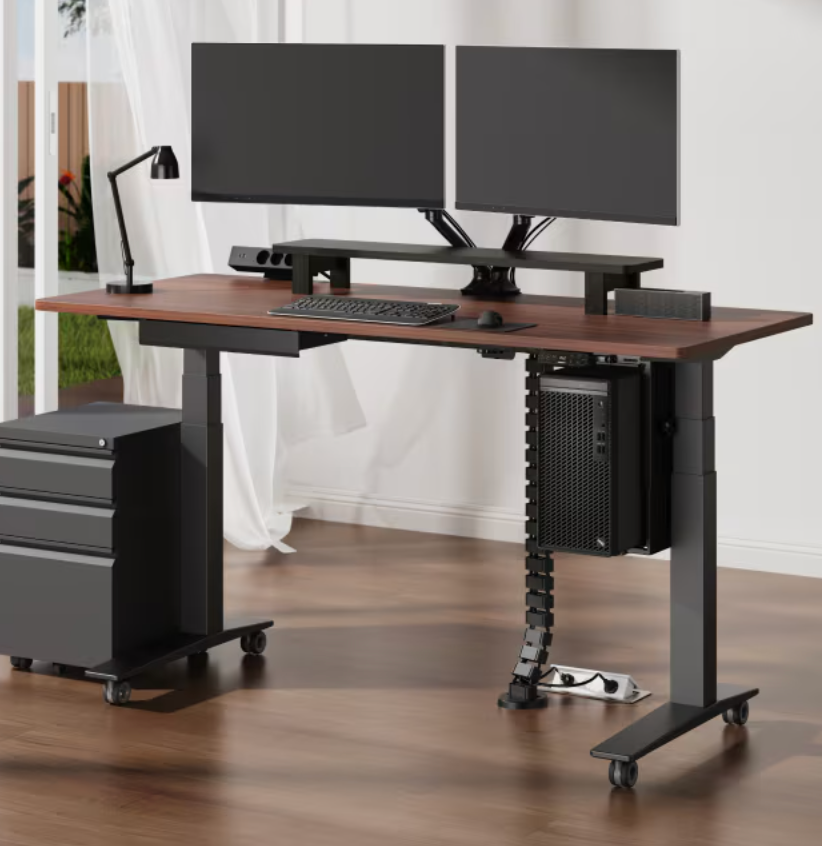 Adjustable standing desk with dual monitors, a keyboard, a lamp, and organized cables. Office chair and small storage unit nearby