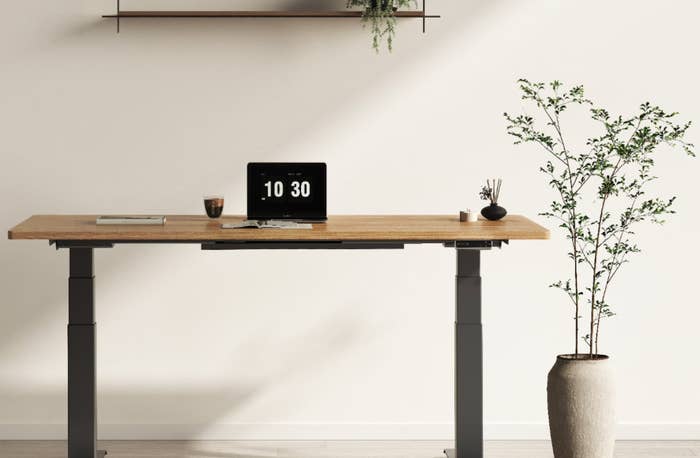 Minimalist workspace with a wooden standing desk, a laptop showing 10:30, coffee cup, notebook, and a potted plant nearby