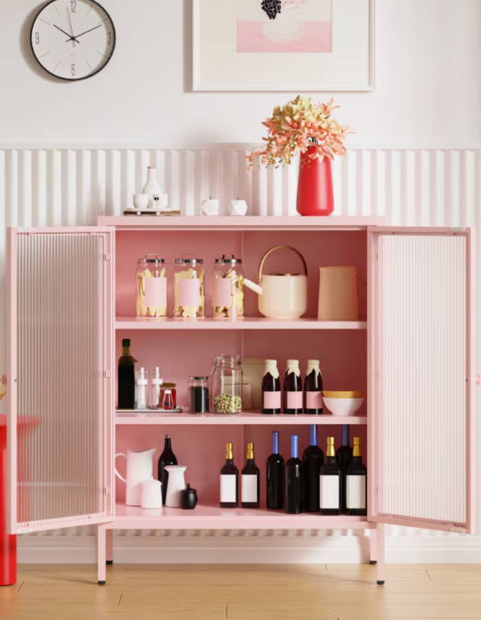 Open pink cabinet with various bottles, jars, and vases. A clock and flower vase are on top. Modern and organized interior decor