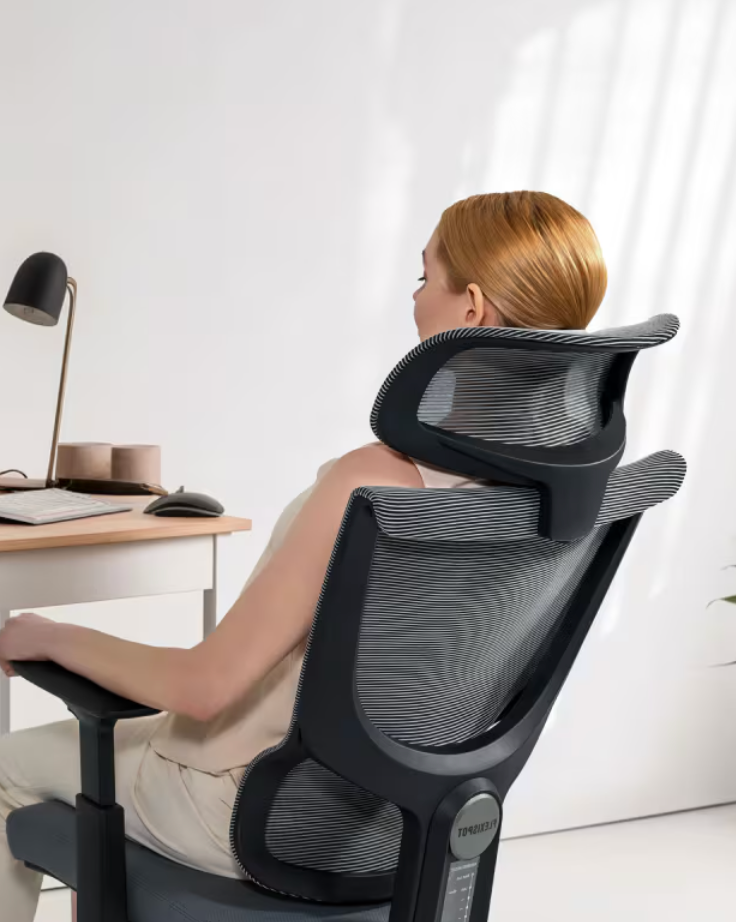 Person sitting in an ergonomic office chair, viewed from behind, in a modern workspace with a desk and lamp