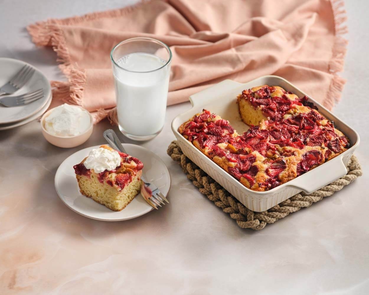 A plate with a slice of strawberry-topped cake beside a glass of milk and a baking dish with more cake