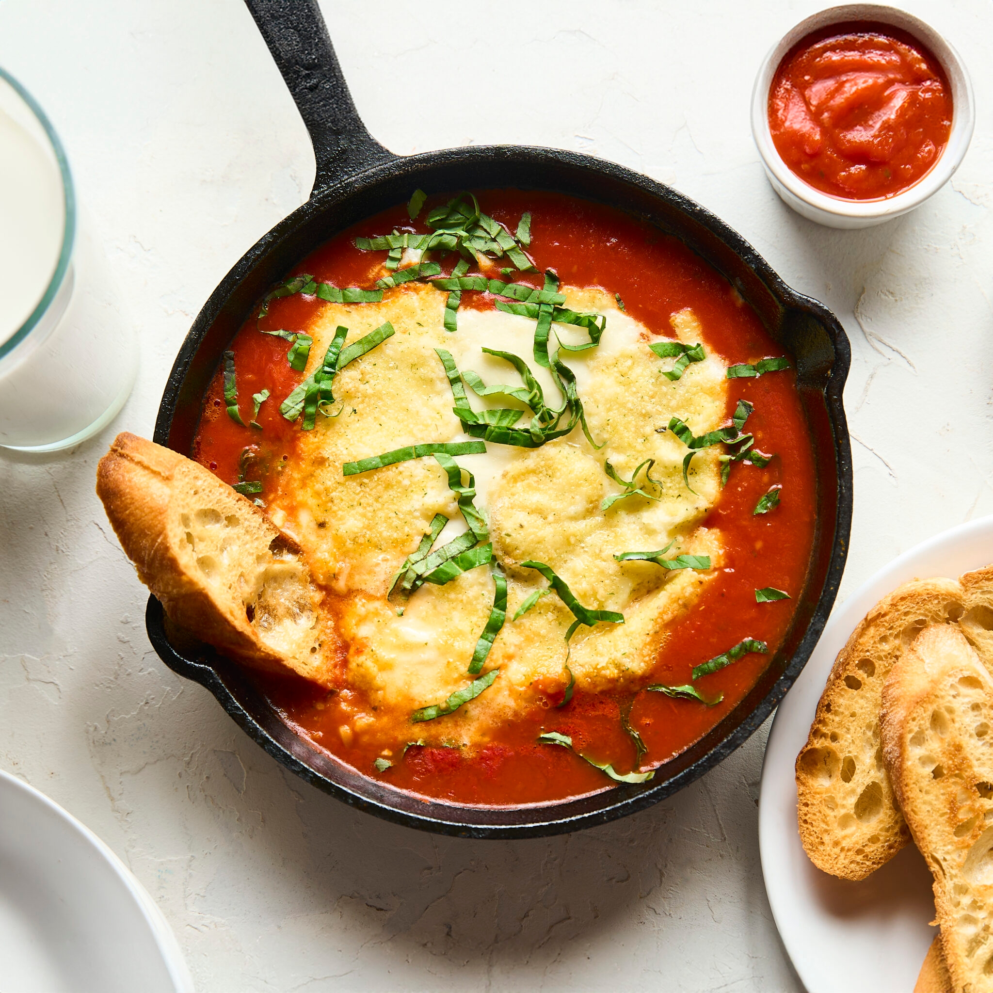 Skillet of baked cheese and tomato sauce topped with basil, alongside sliced bread and a small cup of sauce on a white table