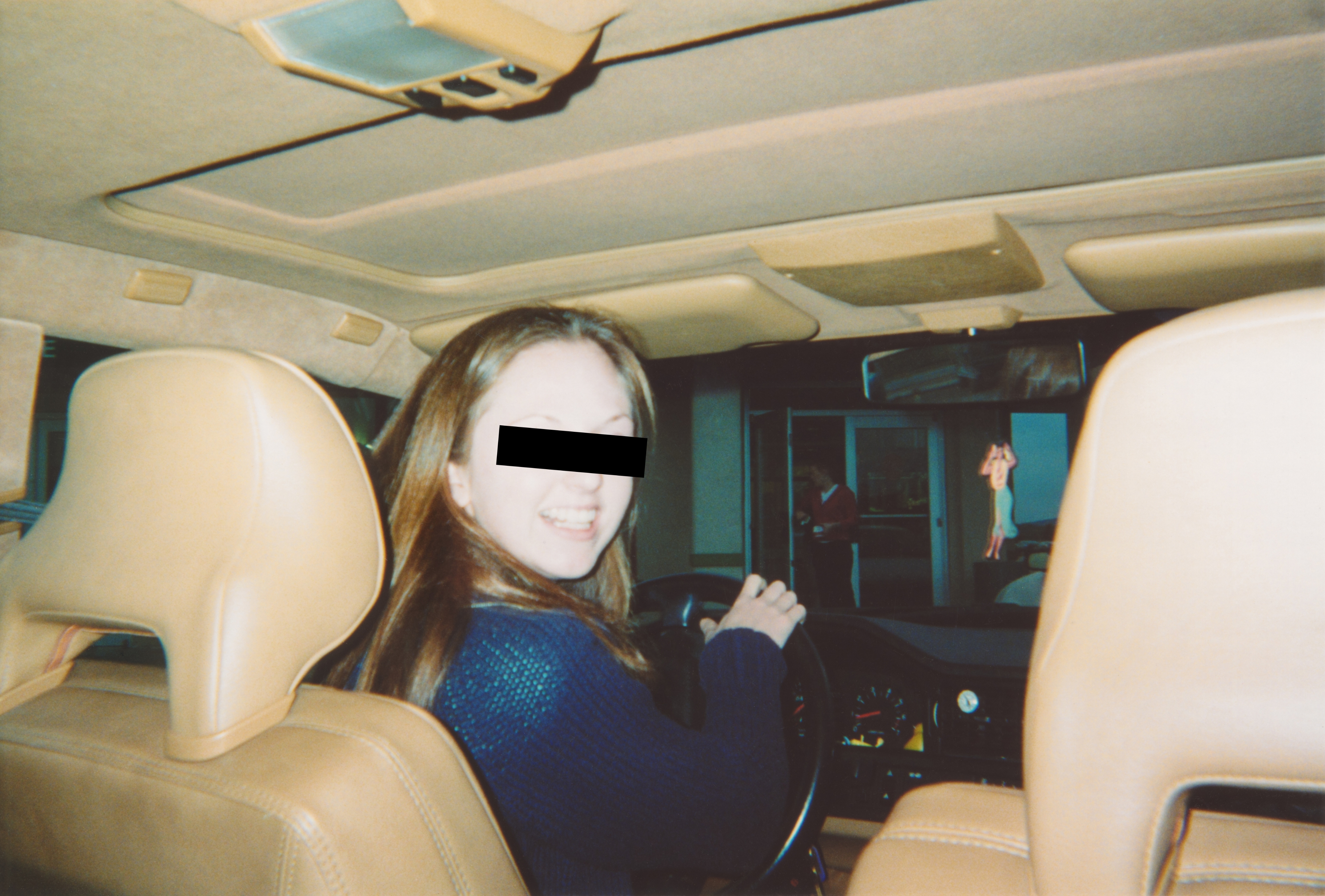 A person sitting in the driver's seat of a car, turning around and smiling at the camera. The car's interior is visible