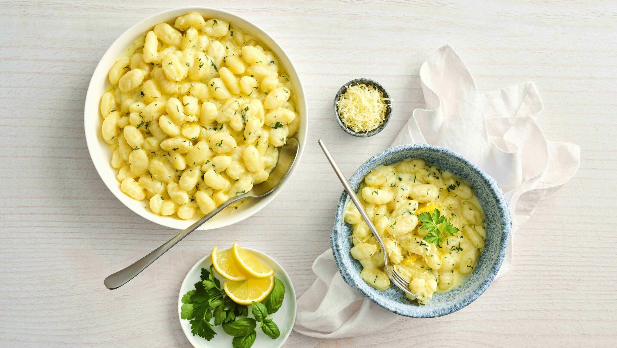 Bowls of creamy gnocchi with herbs and cheese. Garnished with lemon slices and fresh parsley on a white surface