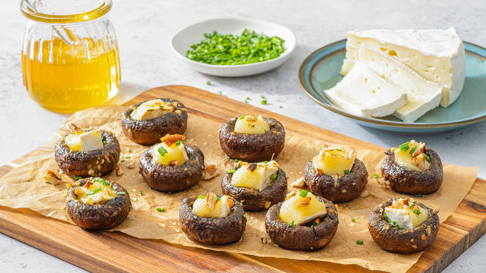 Stuffed mushrooms with melted cheese on a wooden board, honey jar, chopped chives, and brie cheese on a plate in the background