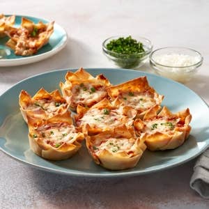 Plate of crispy wonton cups filled with melted cheese and herbs, with two bowls of chives and grated cheese in the background