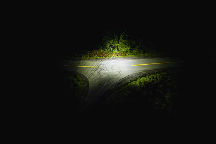 Dimly lit intersection on a road at night with skid marks visible, surrounded by trees