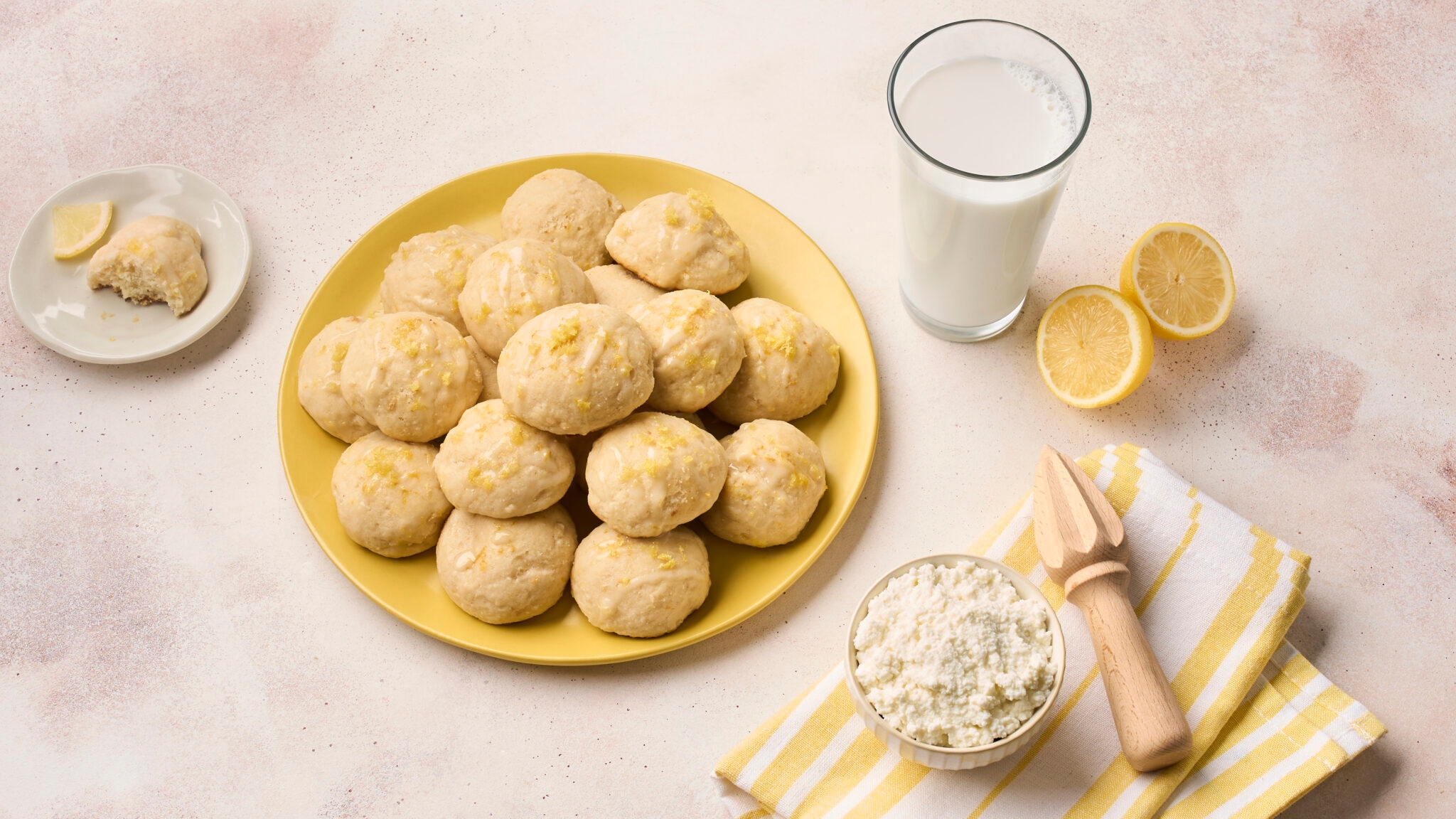Plate of lemon cookies beside a glass of milk, lemon halves, a bowl of ricotta, and a lemon reamer on a striped napkin