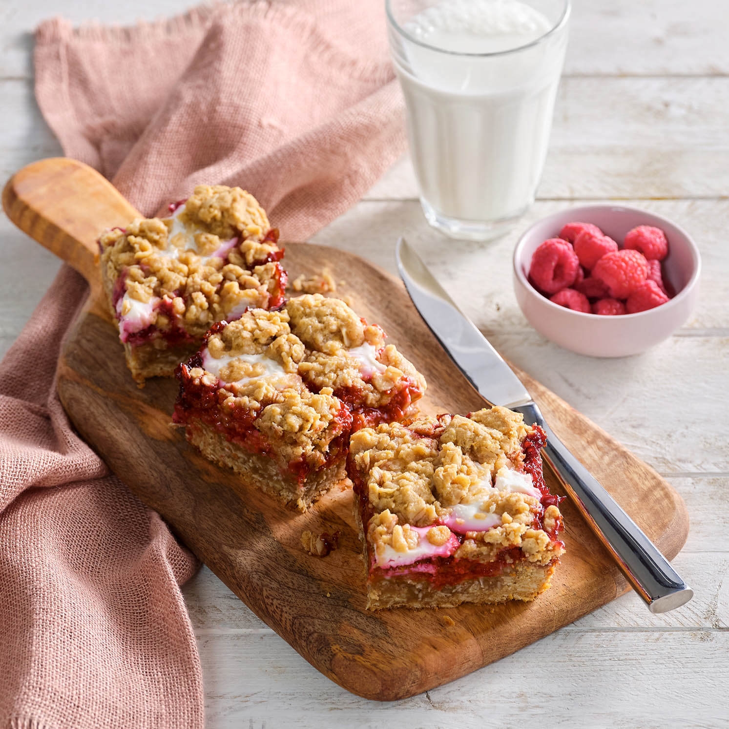 Three raspberry crumble bars on a wooden board with a knife, a bowl of raspberries, and a glass of milk