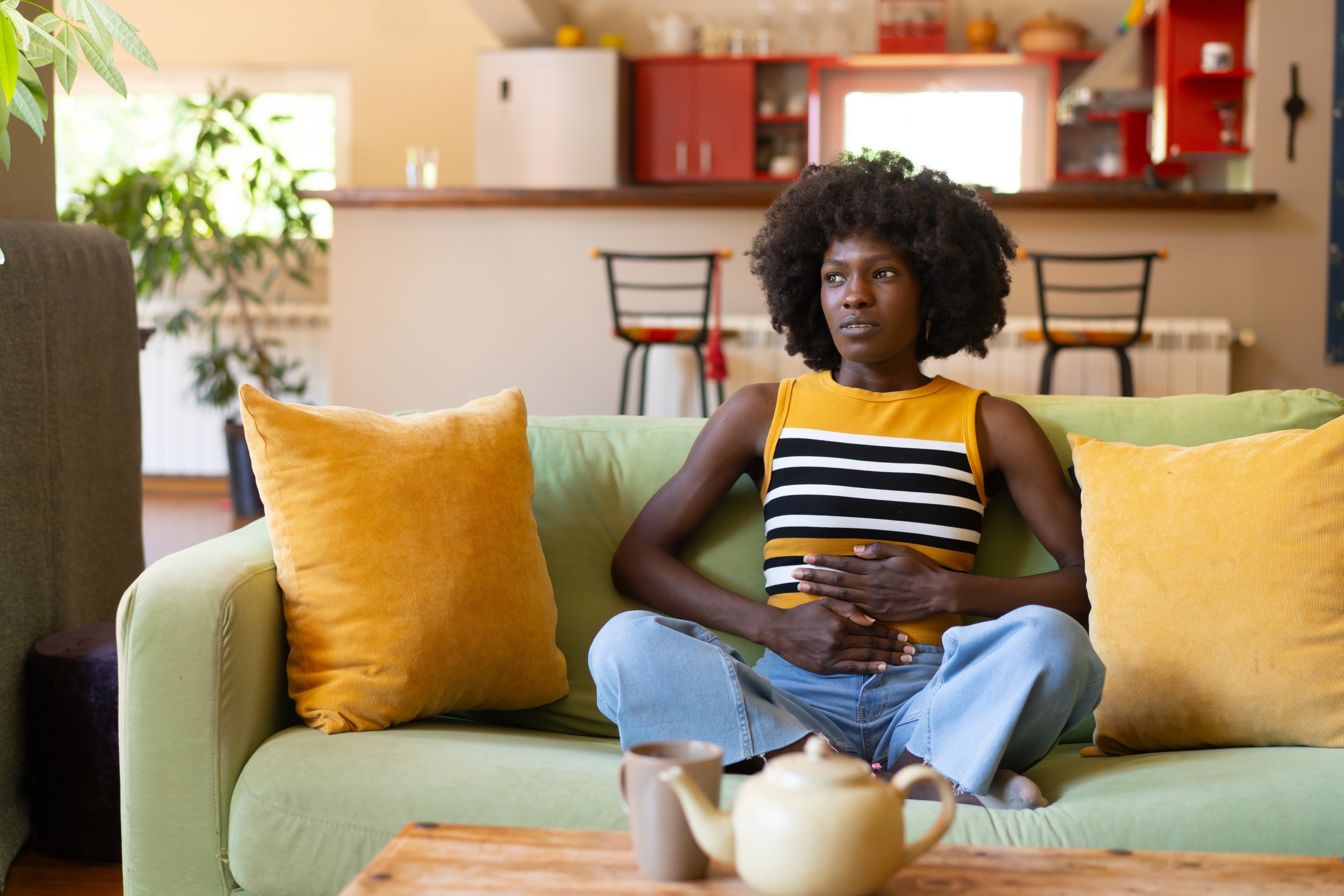 Person sitting on a couch holding their stomach, looking pensive, with a teapot and cups on a table. Kitchen in the background