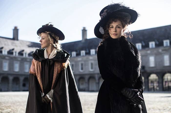 Dos mujeres posan al aire libre con trajes de época, incluyendo grandes sombreros adornados y vestidos elegantes. Están en un entorno histórico