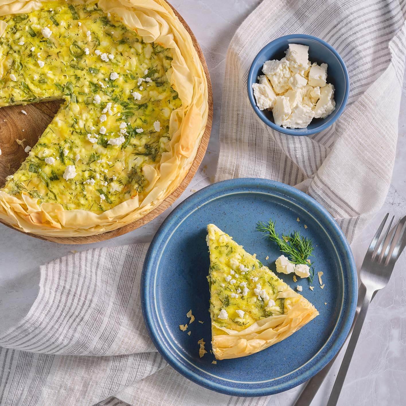 A slice of savory quiche with herbs and feta cheese is served on a plate, with extra feta in a small bowl nearby