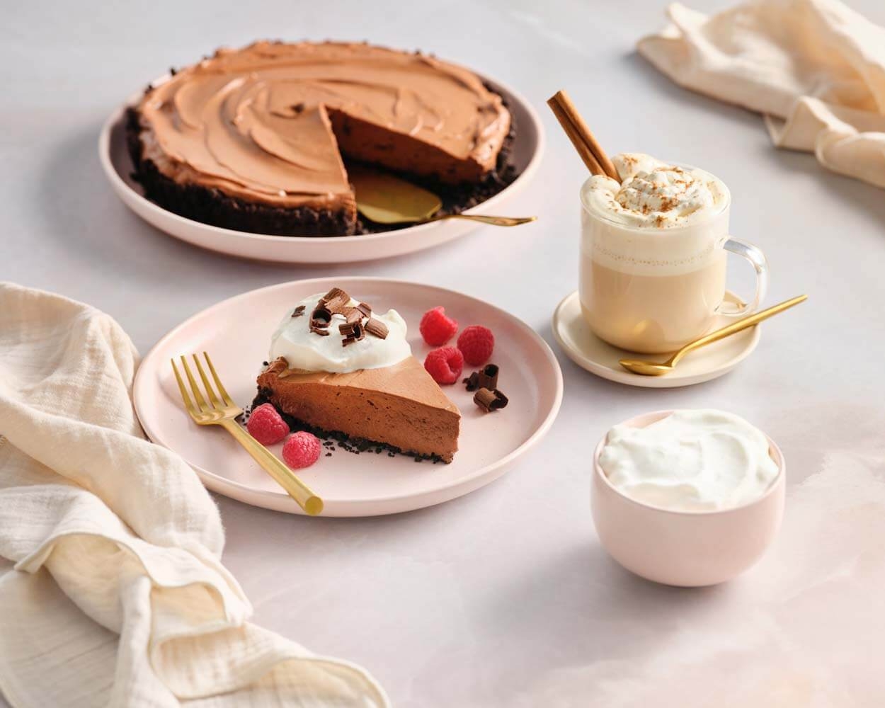 Chocolate pie slice on a pink plate with raspberries and whipped cream. Nearby are a latte with cinnamon stick and a small bowl of whipped cream