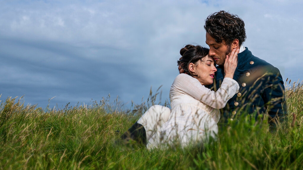 Pareja abrazada en un campo, vestida con ropa de estilo histórico, creando una atmósfera romántica entre el césped alto