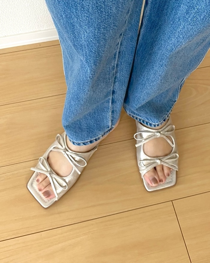 Feet in open-toed sandals with bow details, wearing casual jeans, standing on a wooden floor