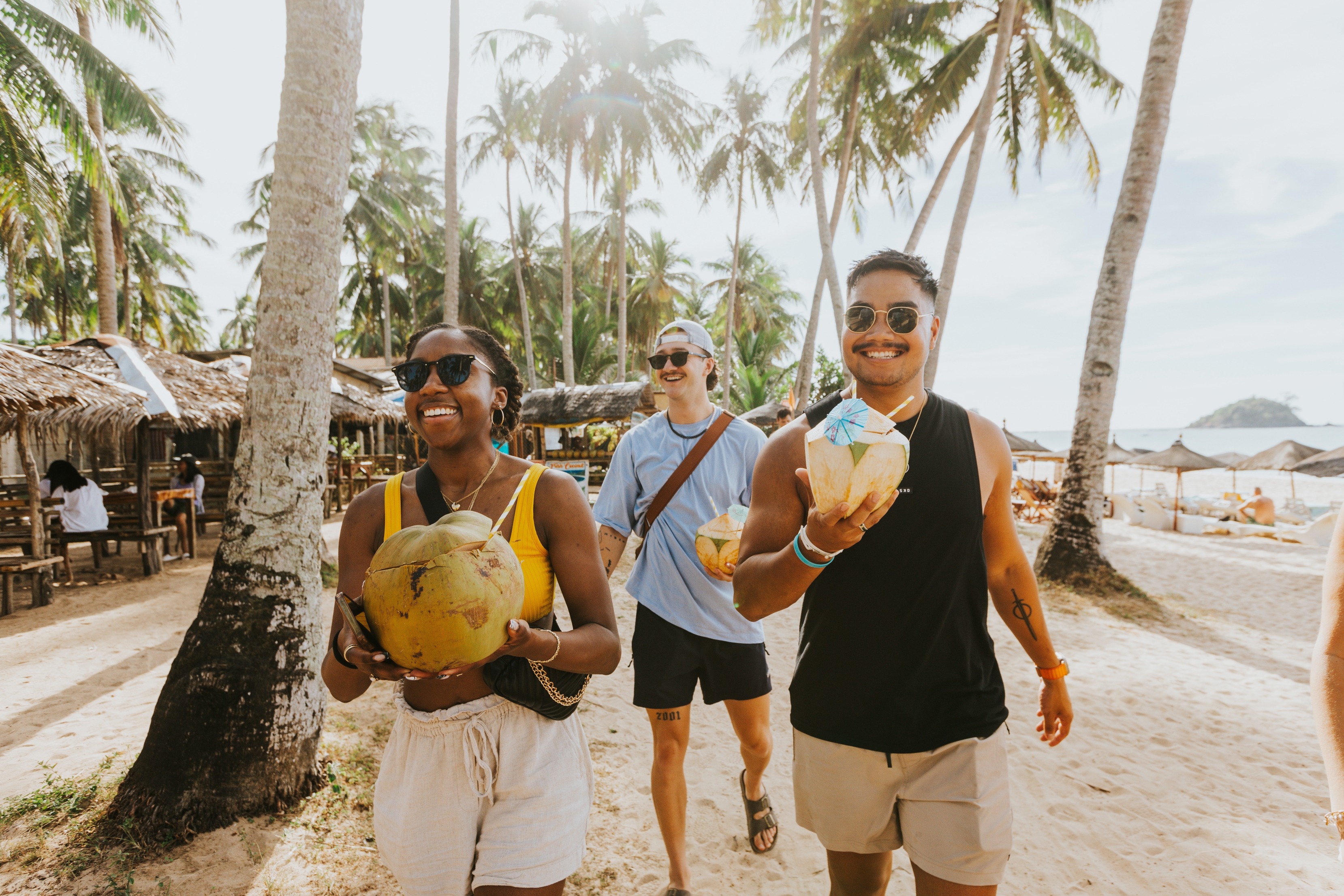 Three people walk on a tropical beach, smiling and holding coconuts. Palm trees and huts are in the background