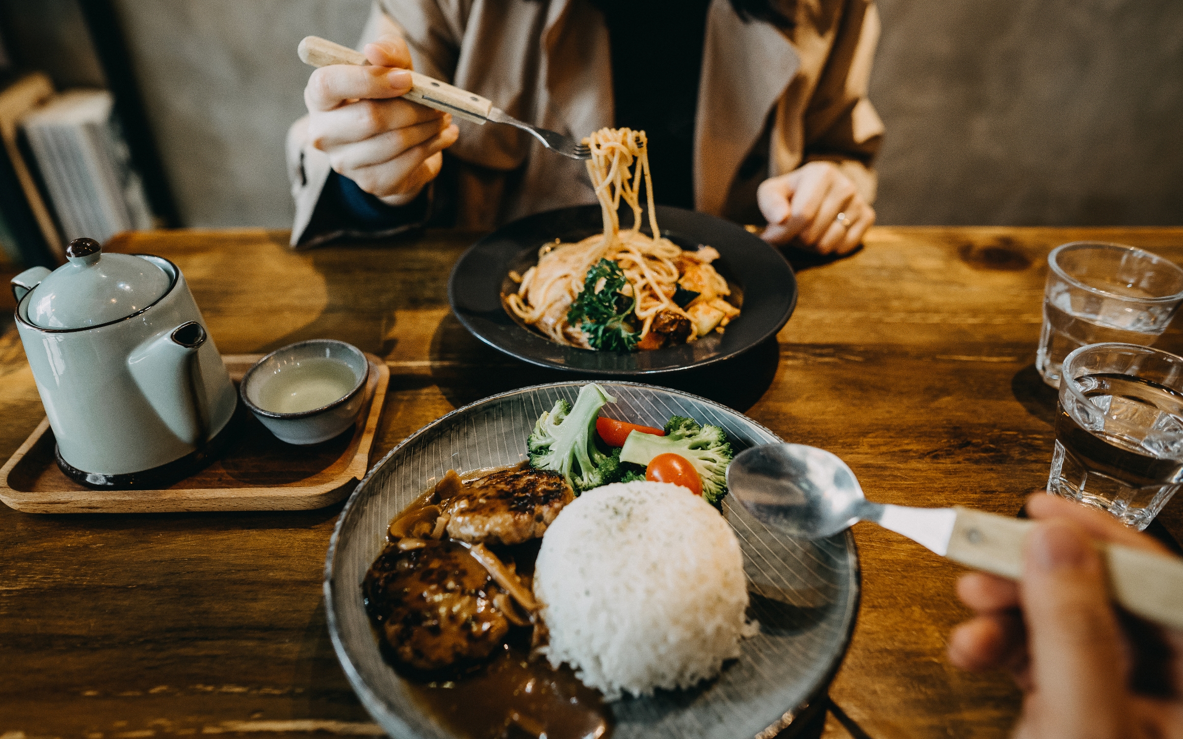 Two people dining with plates of noodles, rice, and meat.