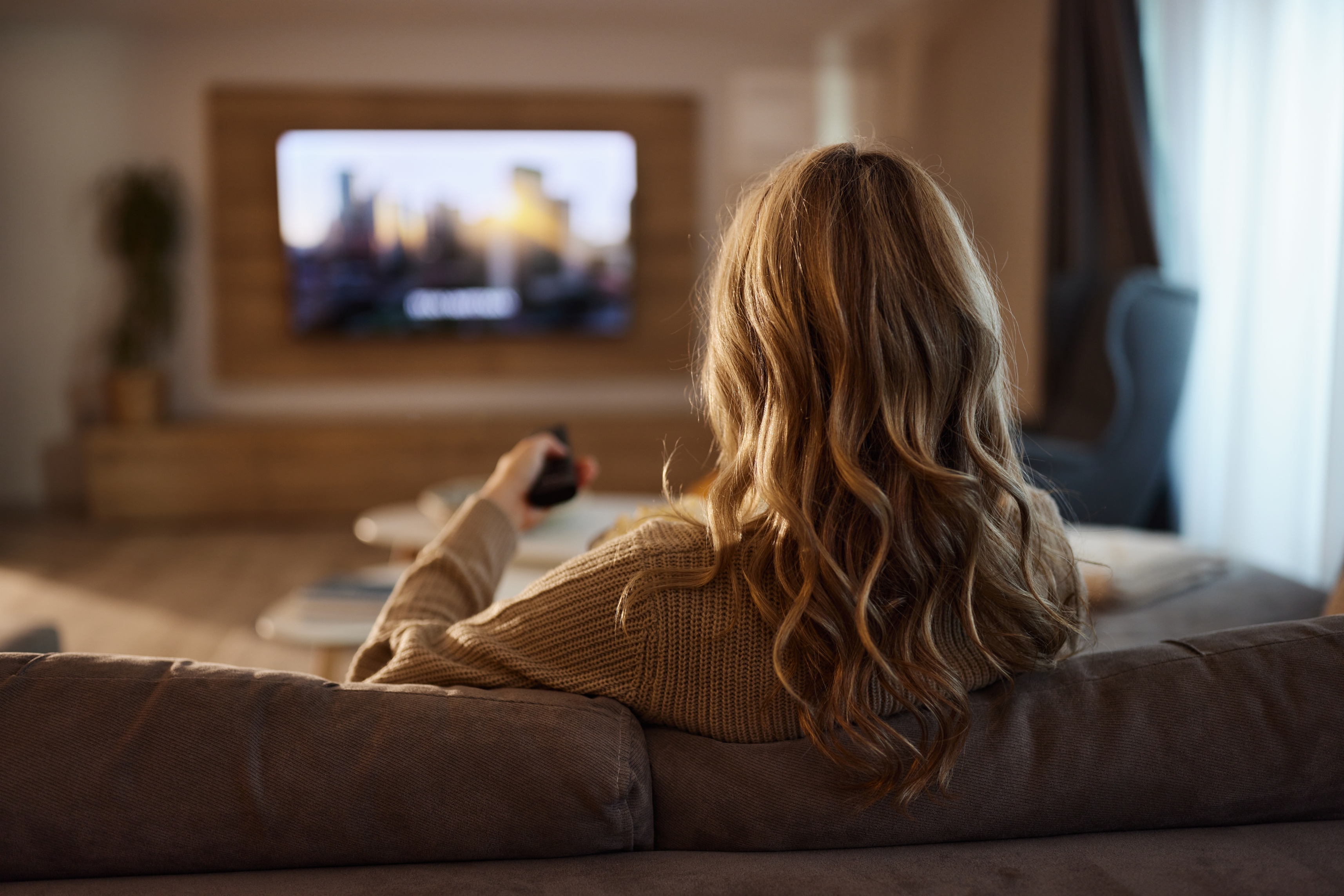 Person with long hair watching TV in a cozy living room, holding a remote. The scene is relaxed and homely, suggesting family-friendly content