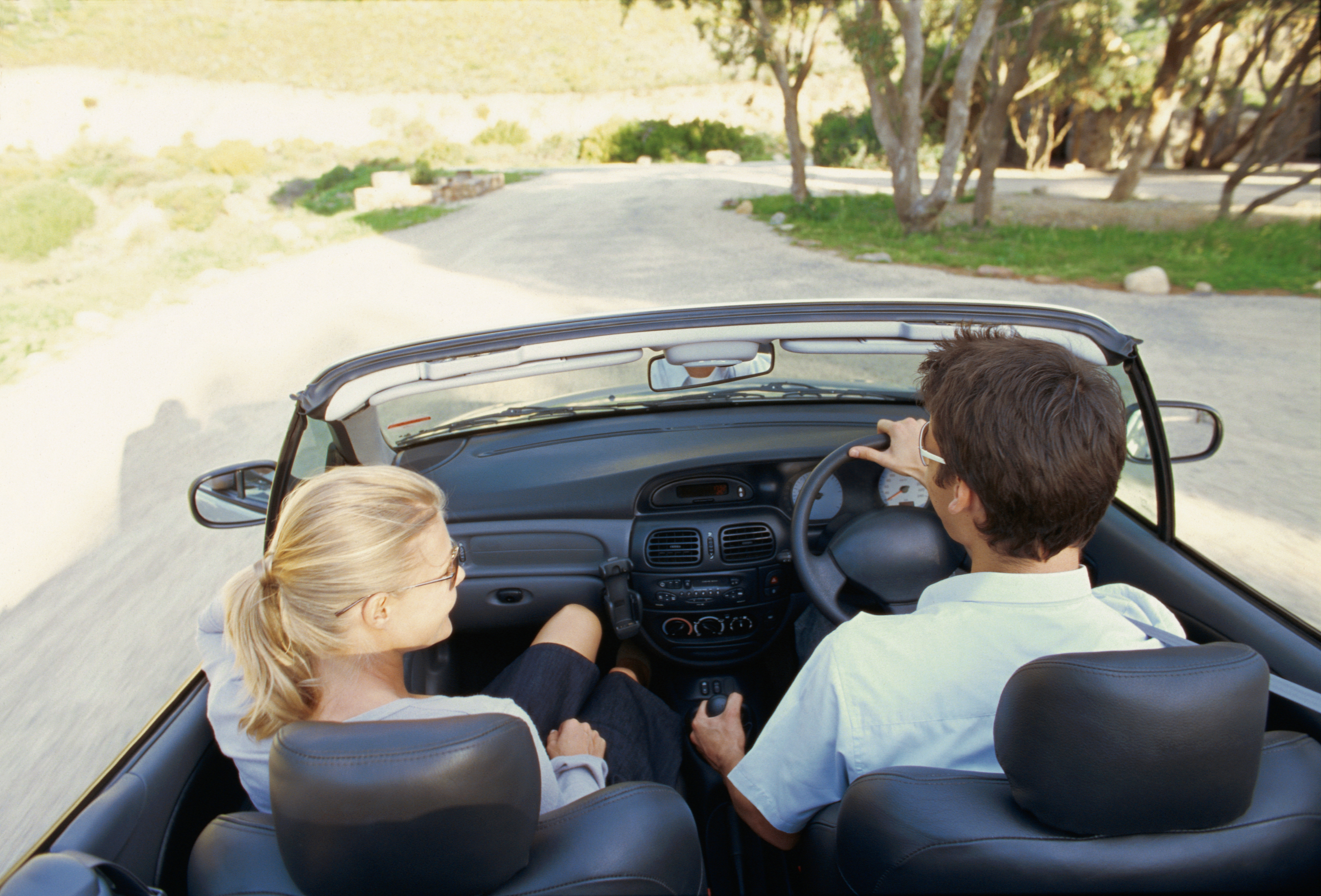 Two people ride in a convertible on a sunny day, driving down a winding road surrounded by trees