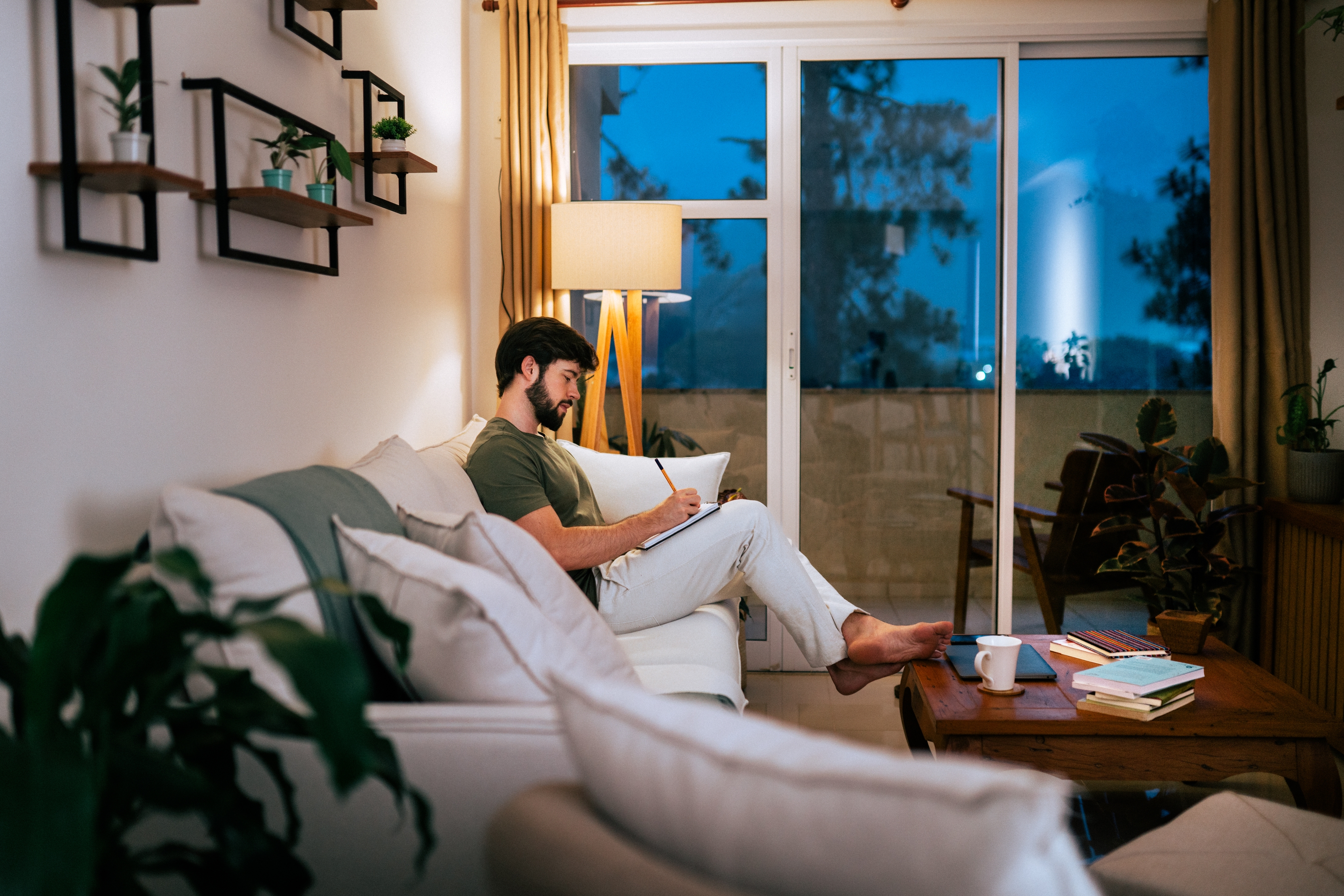 A man relaxes on a sofa, reading a book in a cozy living room with plants and shelves, suggesting a calm evening at home