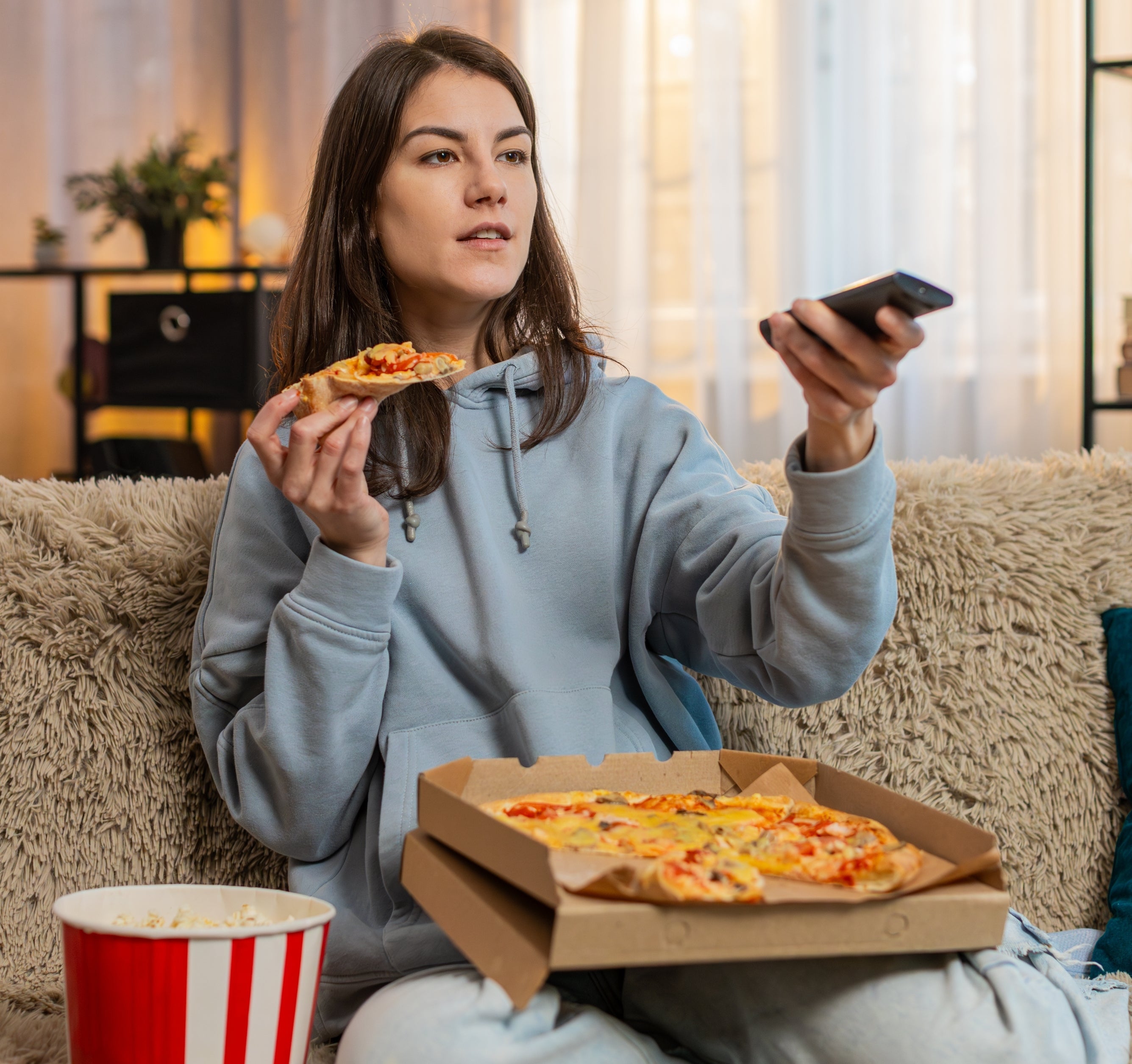 Person in a hoodie enjoys pizza and holds a remote while sitting on a couch at home beside a popcorn bucket