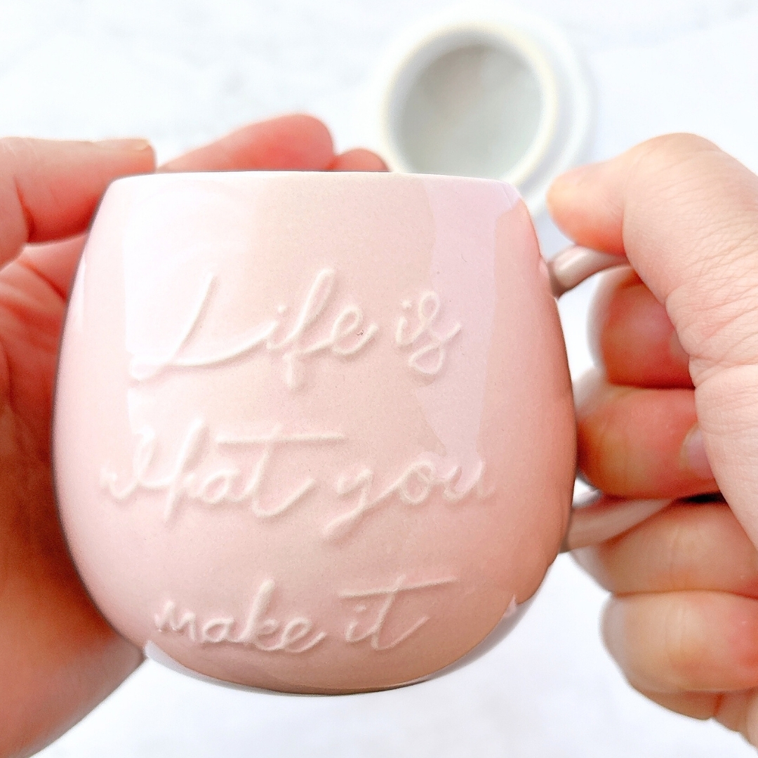 手に持たれたカップに「Life is what you make it」と刻まれている。背景にボケた白い円形オブジェクト。