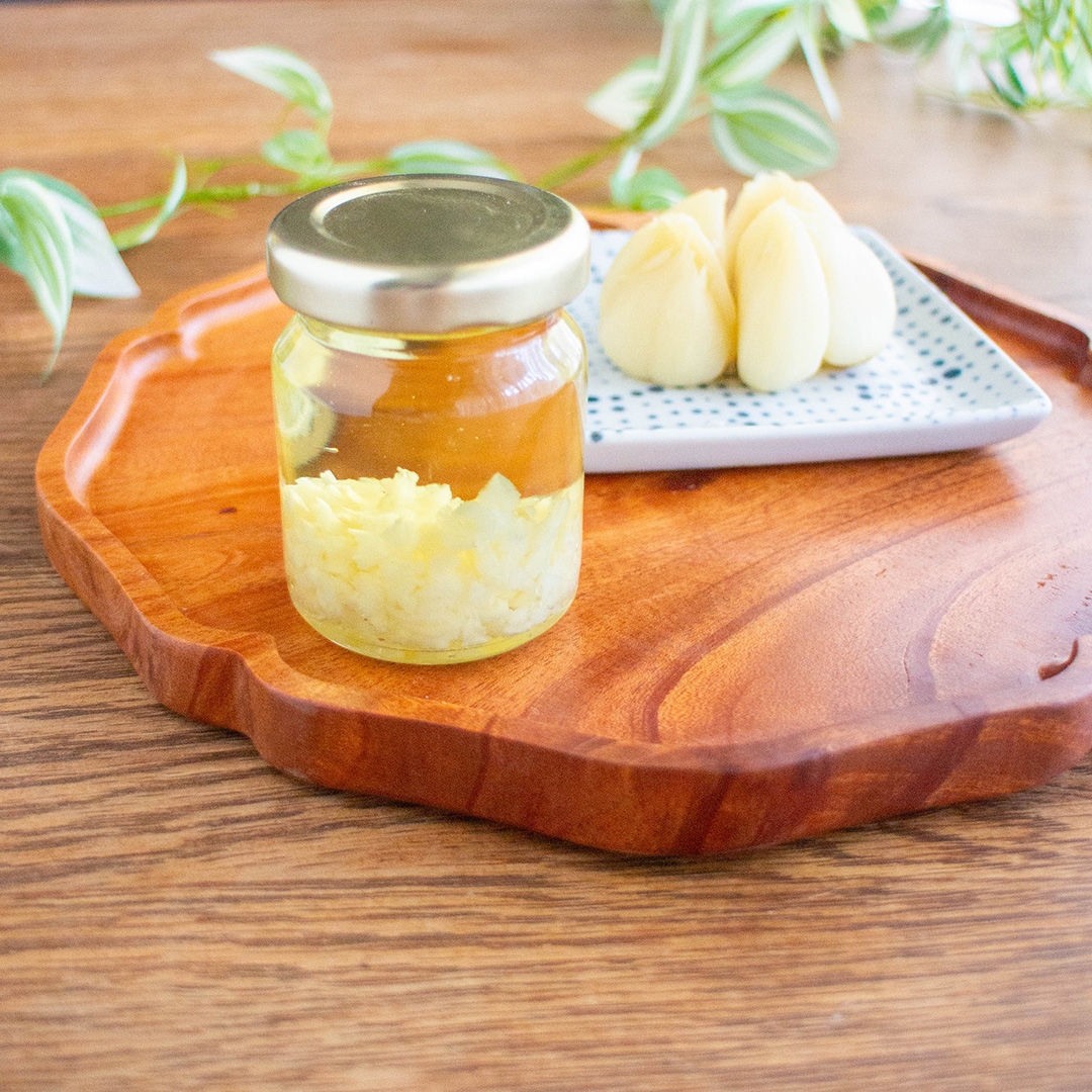 木製トレイに載ったニンニク入りの透明な瓶と、皿に盛られたニンニクの塊。背景に観葉植物。