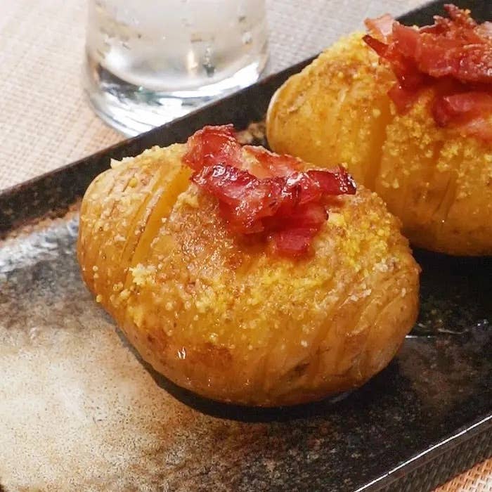 2つのベイクドポテトがベーコンと調味料を載せて皿に盛られている。飲み物のグラスが背景にある。