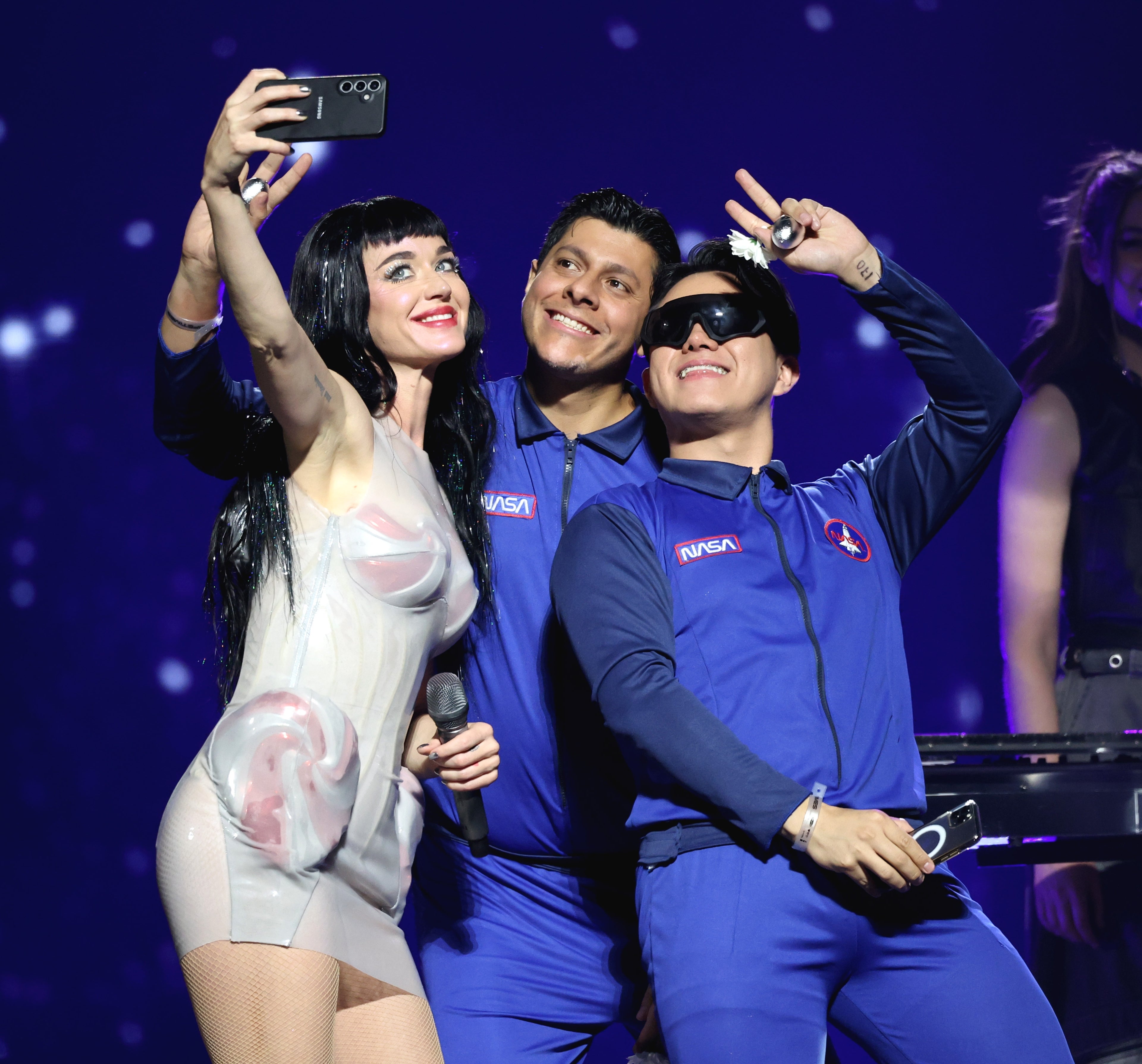 Katy Perry taking a selfie on stage, with two people in NASA-themed outfits