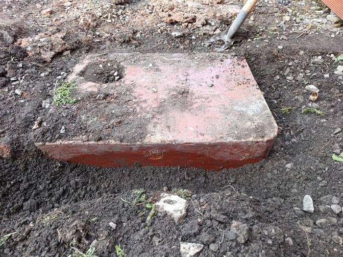 A partially unearthed, large stone block with a carved circle, surrounded by soil and grass patches, with a shovel nearby