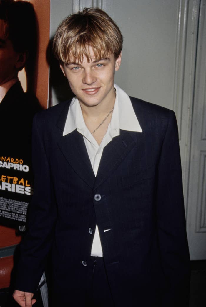 Young man in a sharp suit with an open-collared shirt standing indoors, near a movie poster