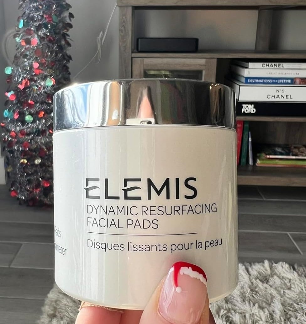 A reviewer's Hand holding Elemis Dynamic Resurfacing Facial Pads with a holiday-themed background, including a tree and decor, in a cozy living room