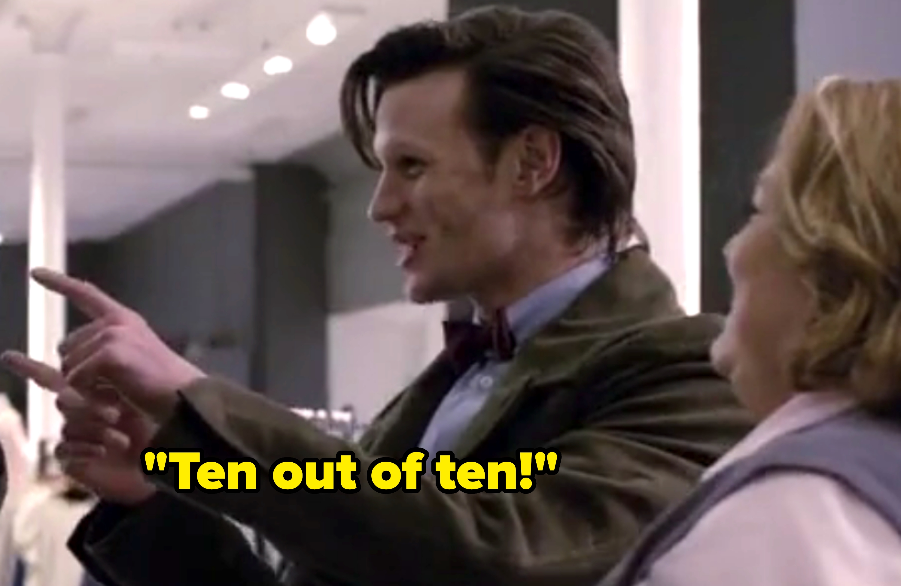 A person in a suit with a bow tie is smiling and pointing, standing beside another person in a store setting