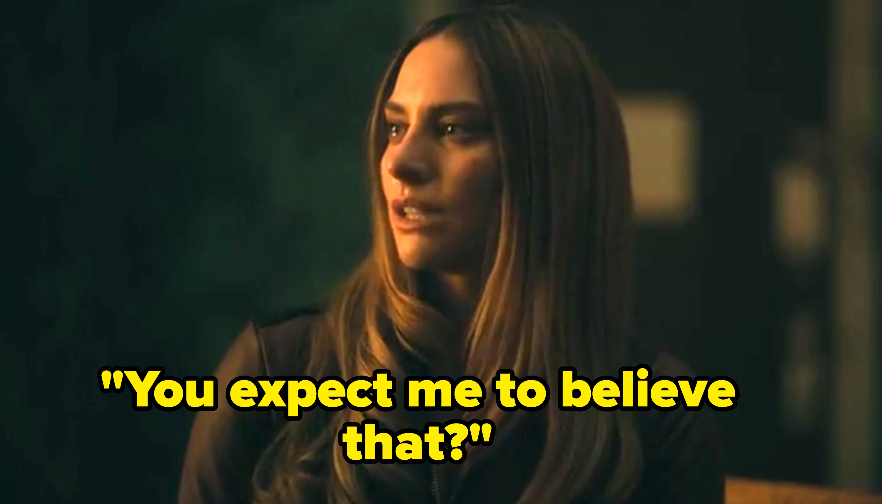 A person with long hair looks thoughtfully to the side, indoors, wearing a dark jacket