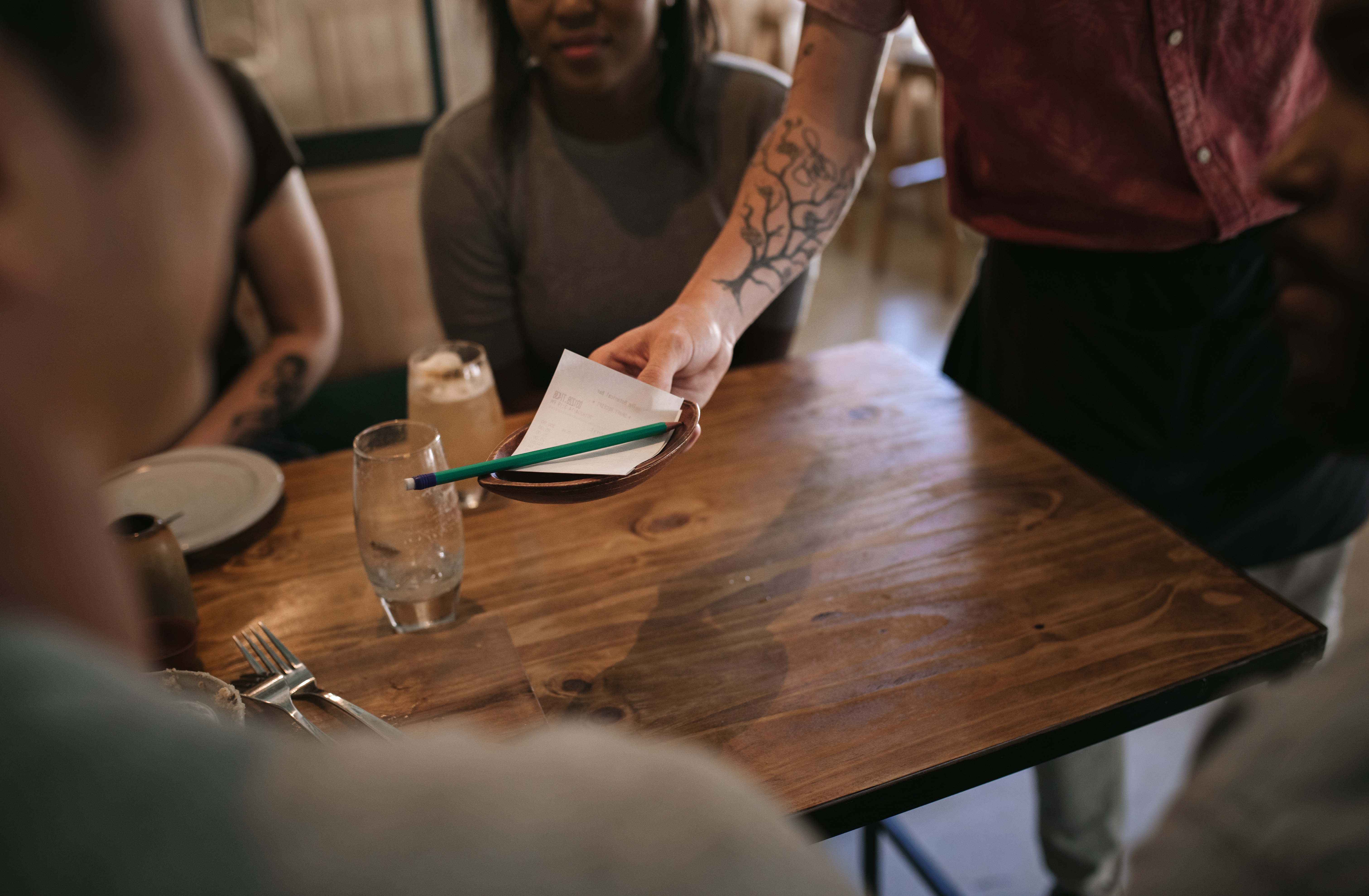 Server handing a bill to seated diners at a restaurant. One person is holding a pen ready to sign. The setting is casual and relaxed