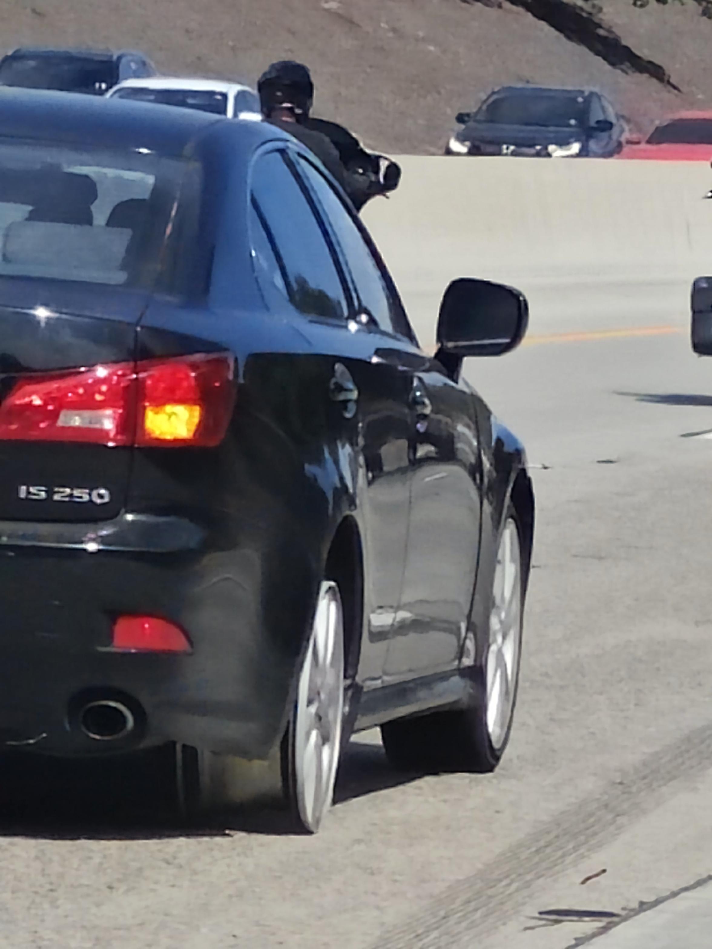 A black sedan driving on a highway, visible with other cars in the background and a motorcycle in the adjacent lane