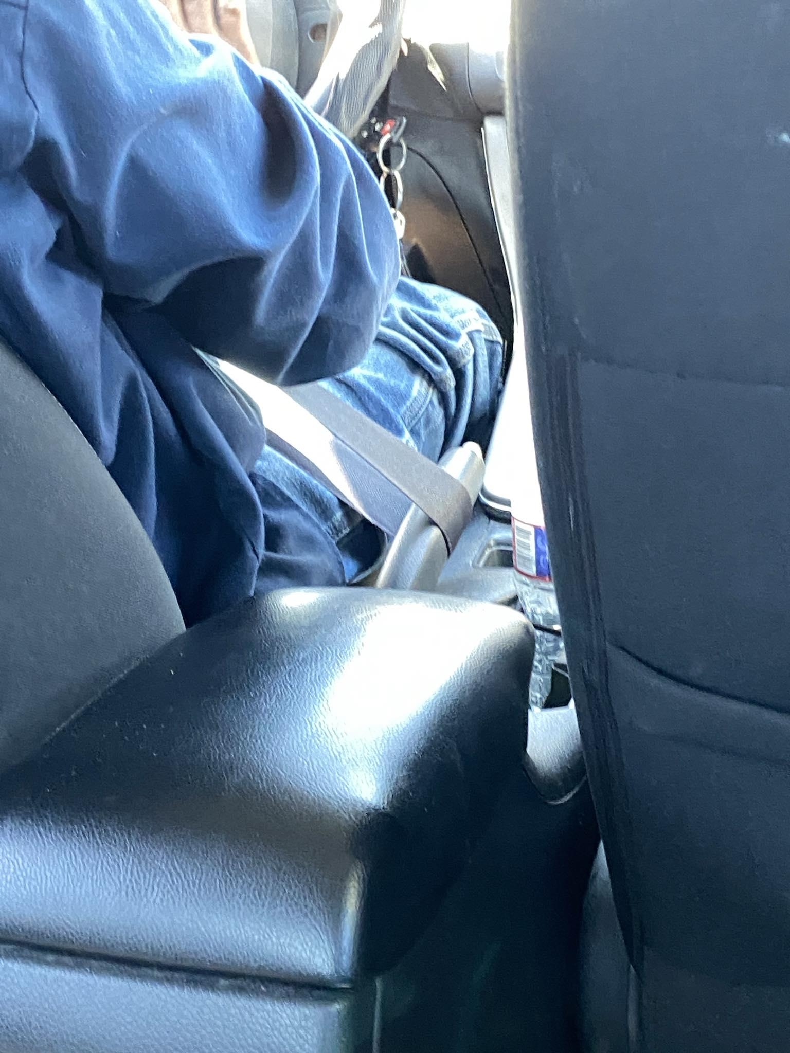 A person in casual attire, seated in the passenger seat of a car, wearing a seatbelt. Car interior is visible with a bottle in the cup holder
