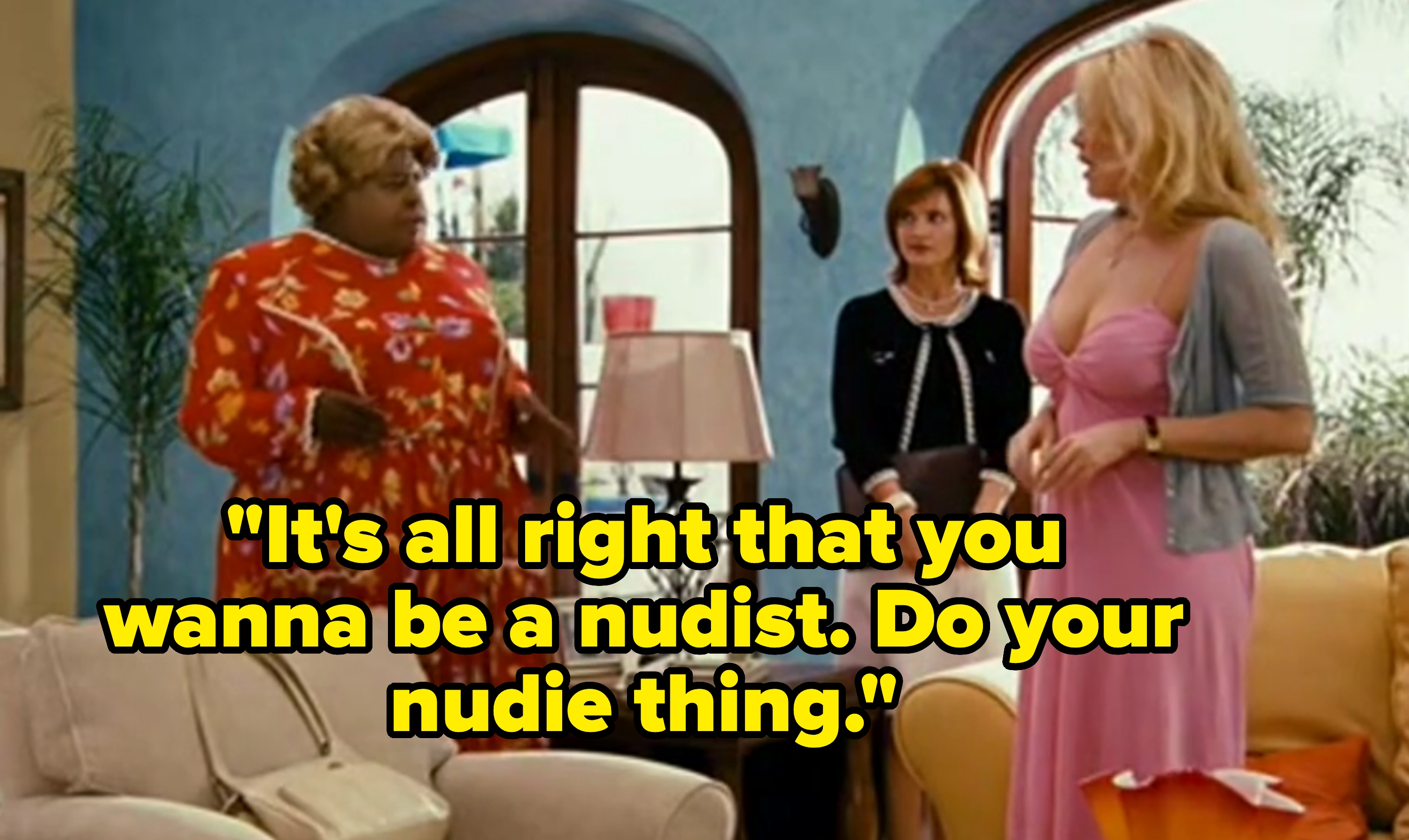 Three people are in a living room, two women standing and one dressed as an elderly woman in a red floral outfit, possibly a comedic scene