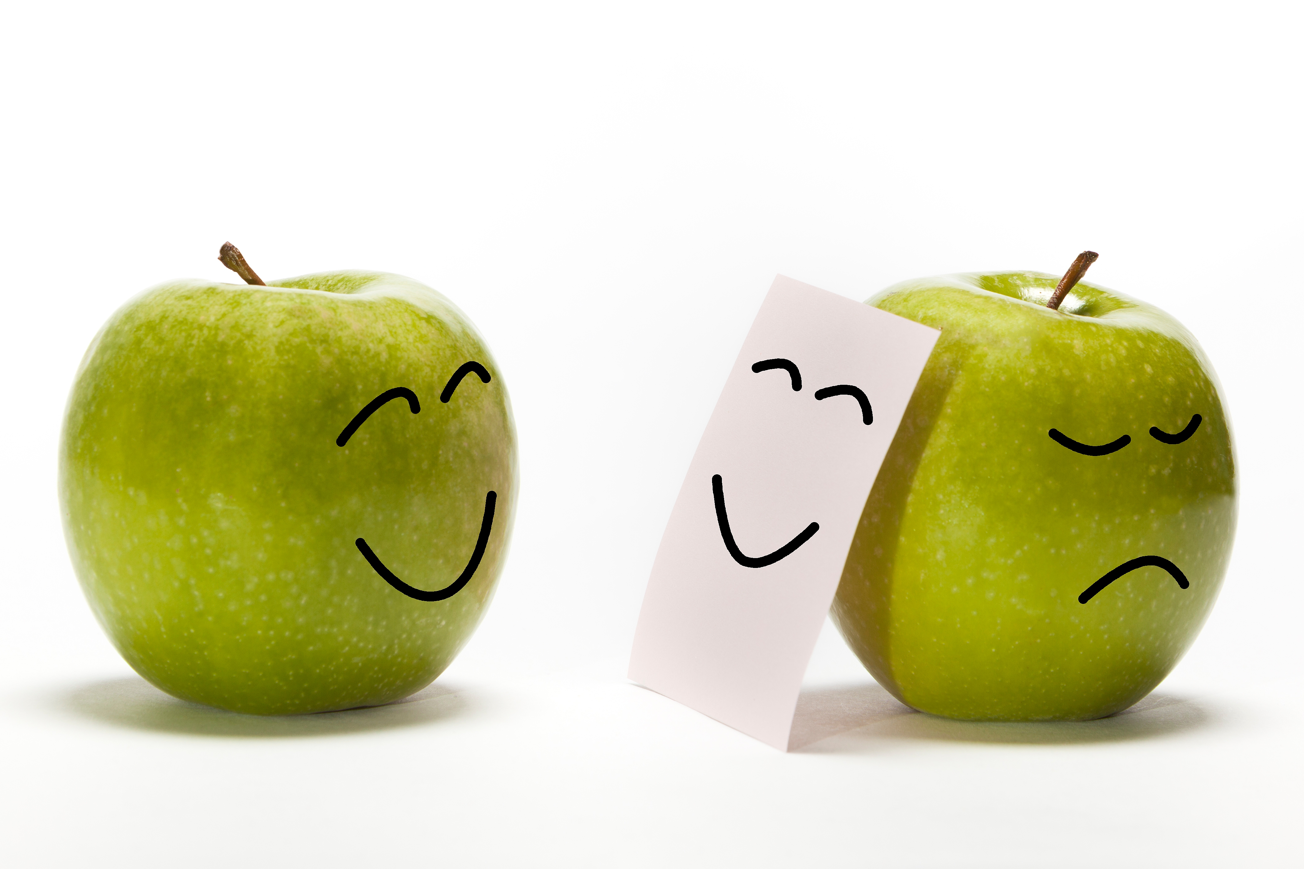 Two apples with faces; one happy, one sad. The sad apple hides behind a paper with a smiling face drawn on it