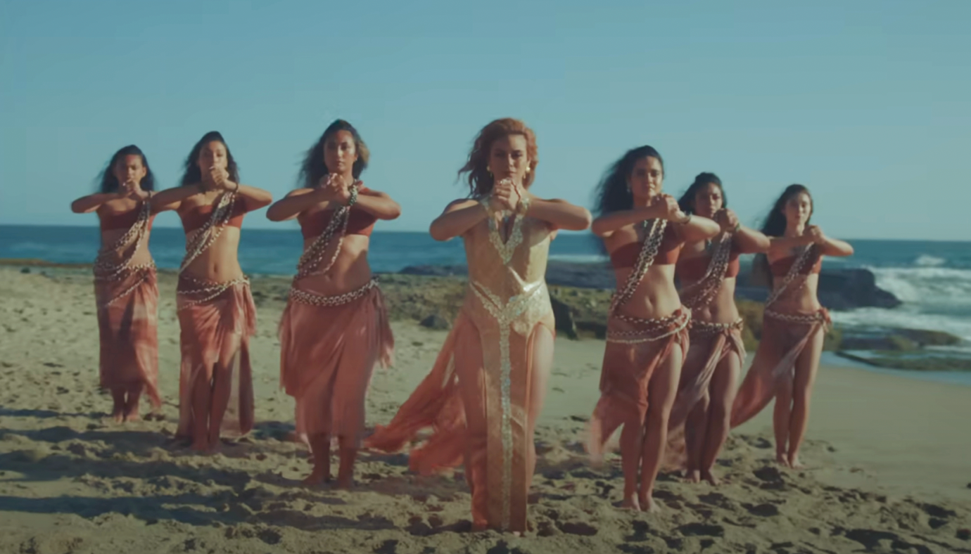 A group of dancers, led by one in a central position, perform a synchronized routine on a beach, wearing flowing, coordinated outfits