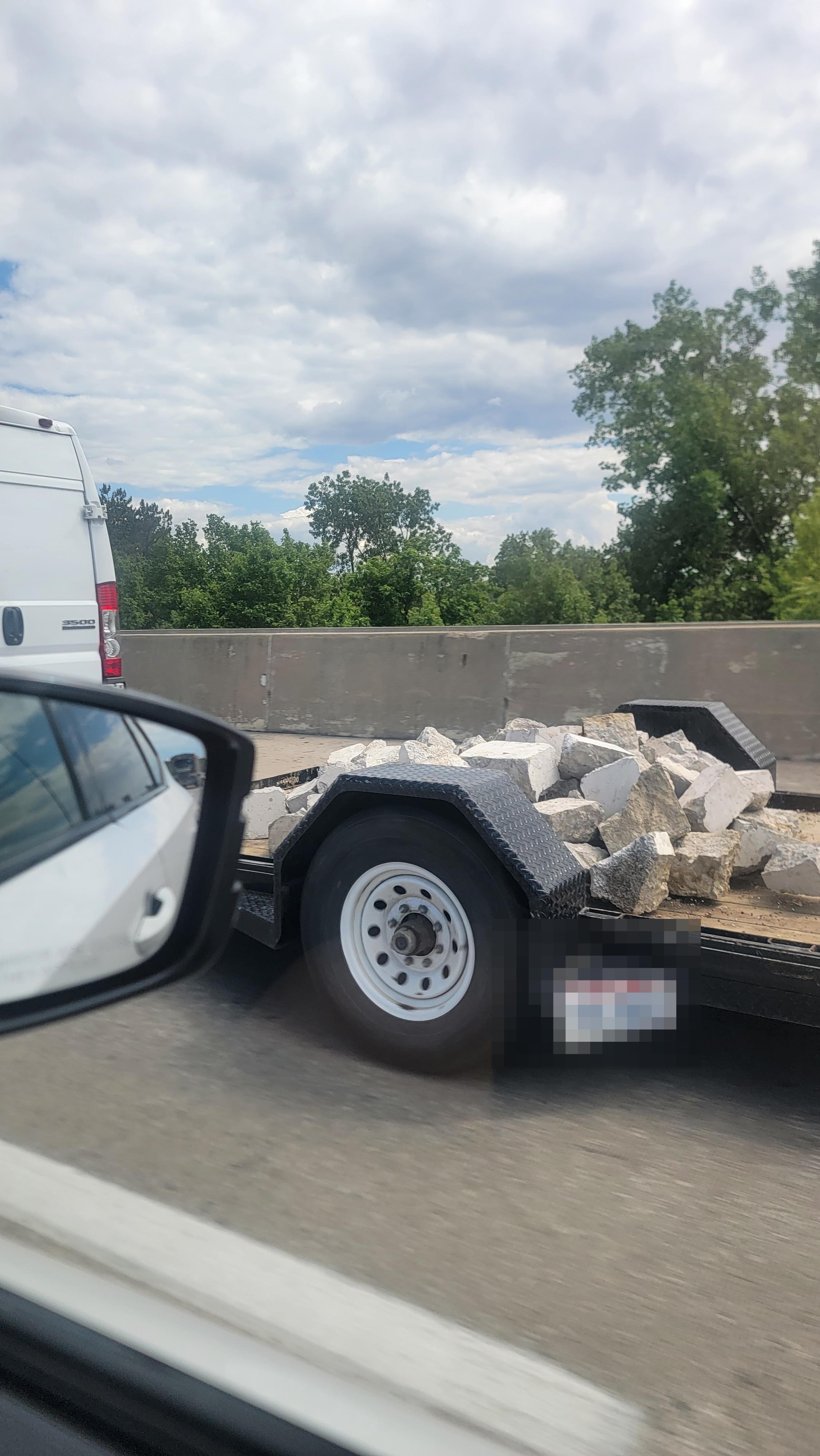 Trailer carrying scattered rocks on a highway, viewed from a car's passenger window. Trees and concrete barrier in background