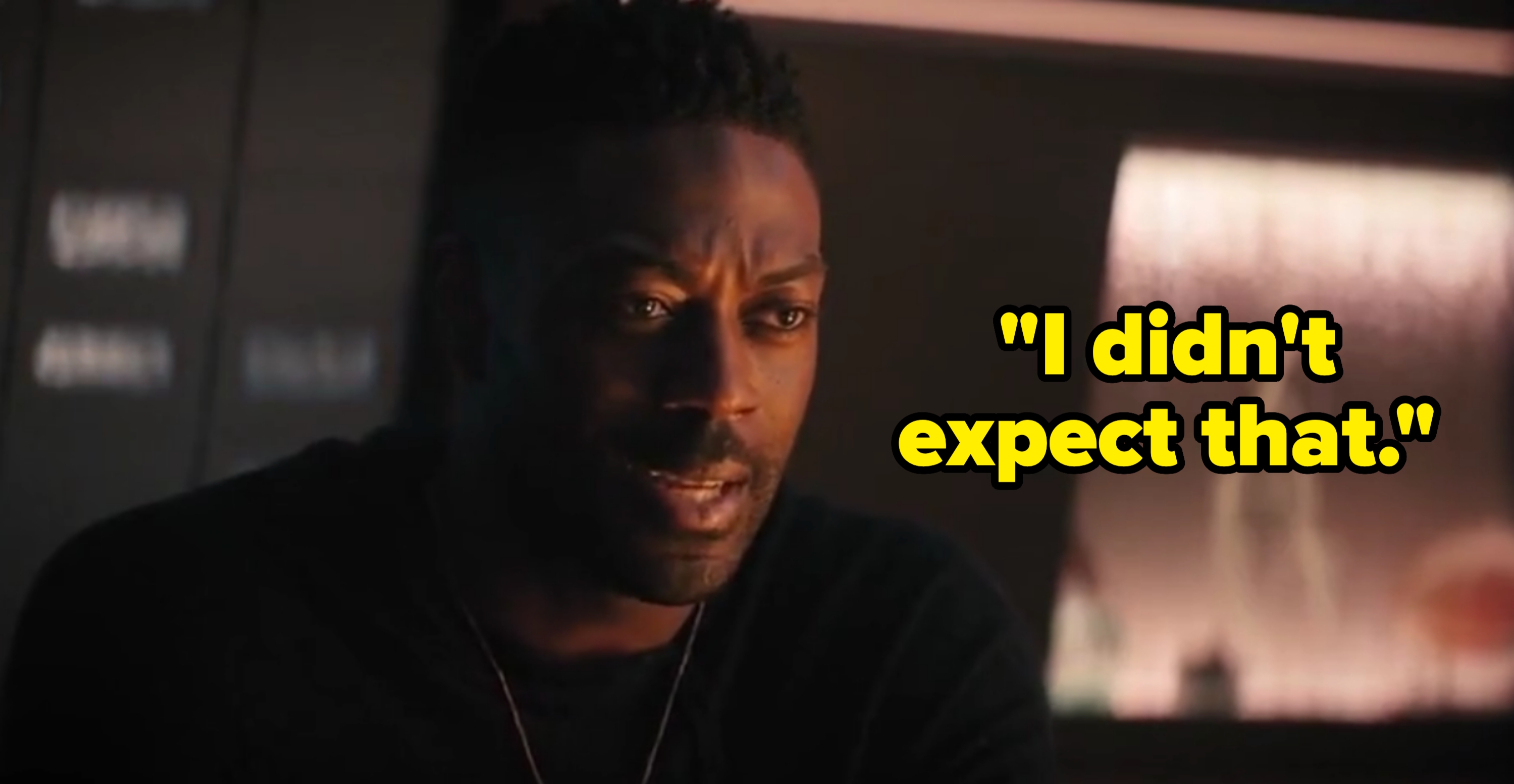 Man in a dimly lit room speaking with a serious expression, wearing a dark shirt