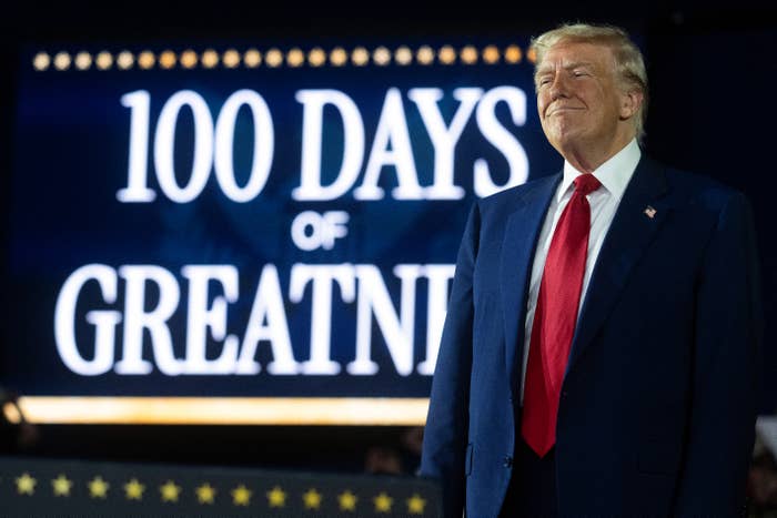A man in a suit stands in front of a large sign reading "100 Days of Greatness" during a public event