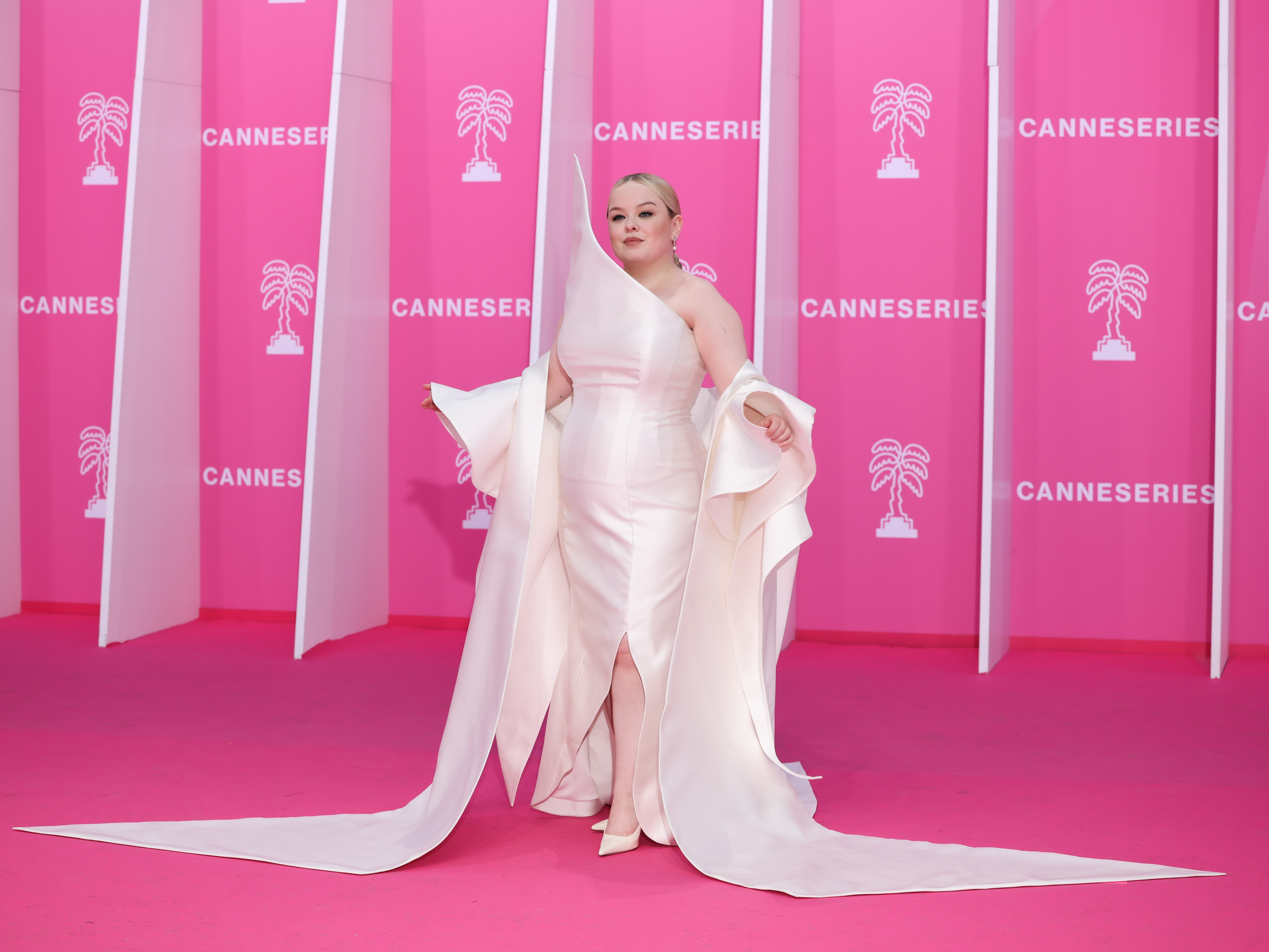 Person in an elegant flowing gown poses at a Cannes event with pink backdrop marked &quot;CANNESERIES.&quot;