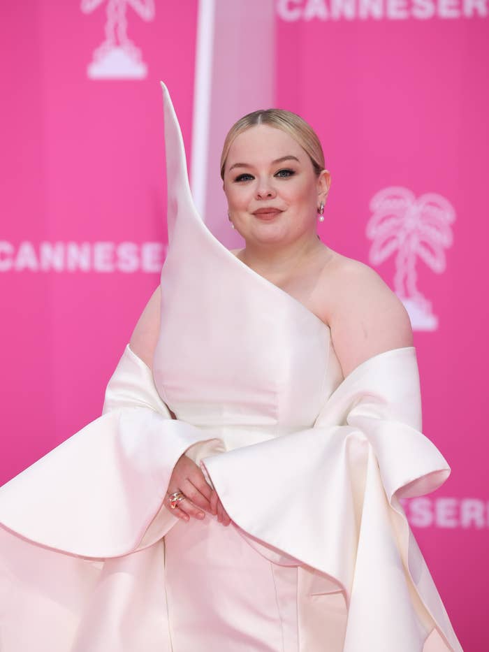 Person in elegant, sculptural white gown poses on a pink carpet with a Cannes backdrop. Hair is styled in a sleek updo