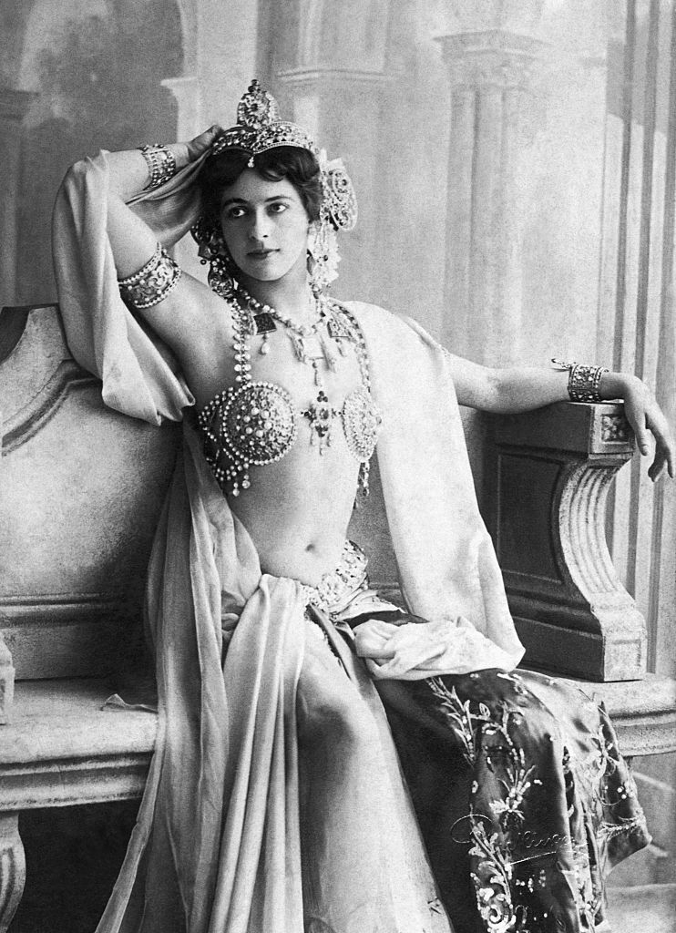 A performer in elaborate, bejeweled attire with a headpiece, seated in a poised, theatrical pose, evoking early 20th-century exotic dance style