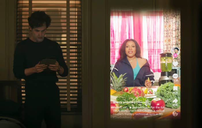 A person stands in a dimly lit room holding a phone, while a video of another person cooking with vegetables plays on a screen beside them