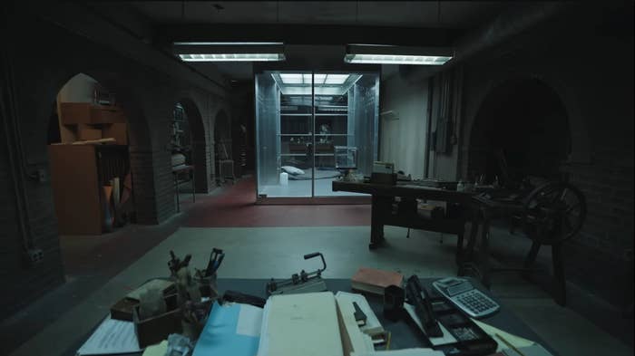 A dimly lit room with a transparent enclosure containing a bed and chair, viewed from behind a cluttered desk with papers and electronic devices