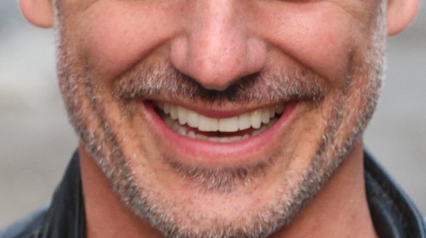 Man with short hair and a salt-and-pepper beard smiles while wearing a leather jacket in an urban alleyway