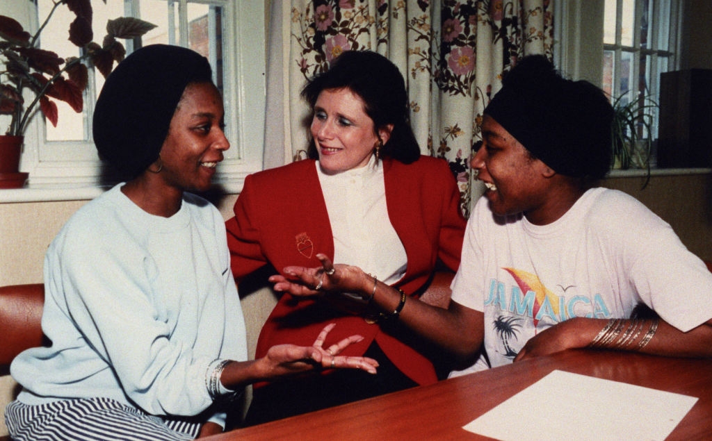 Three people are talking and smiling at a table. The person in the middle is dressed in a blazer, while the others wear casual attire